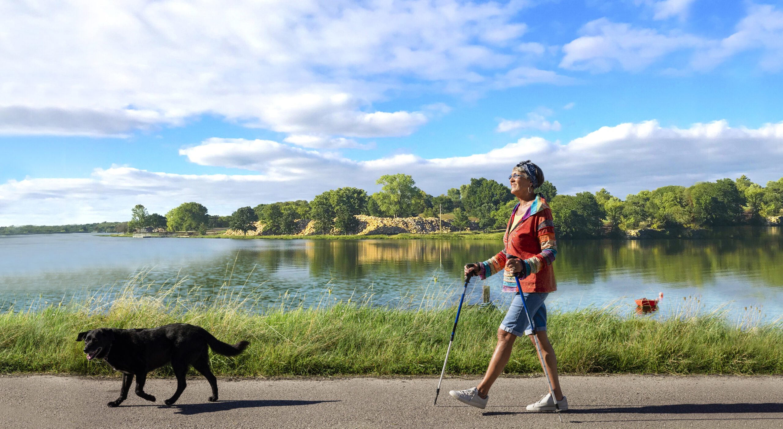 A side-view shot of a senior woman walking by a lake in the Midwest. She is using a hiking pole and enjoying the nice weather walking with her dog.